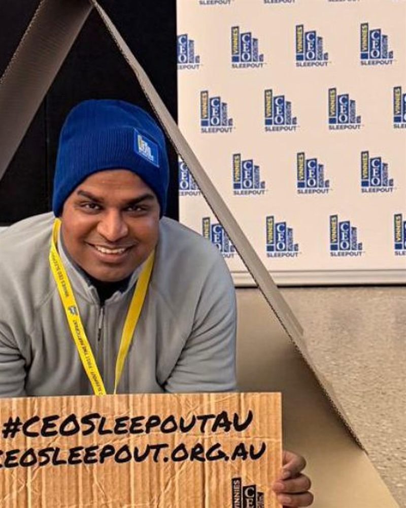 A man in a beanie and lanyard sitting inside a cardboard shelter, holding a hand-lettered CEO Sleepout sign