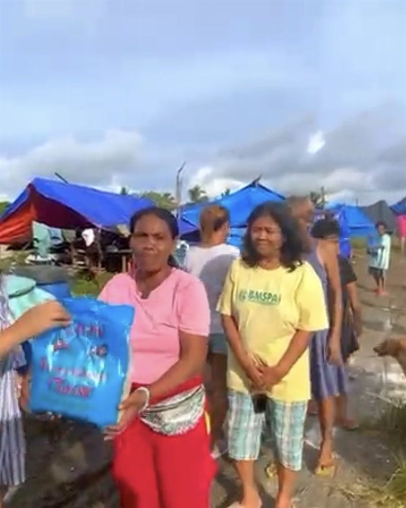 A woman holding a blue relief package, standing with neighbours outside tarpaulin shelters