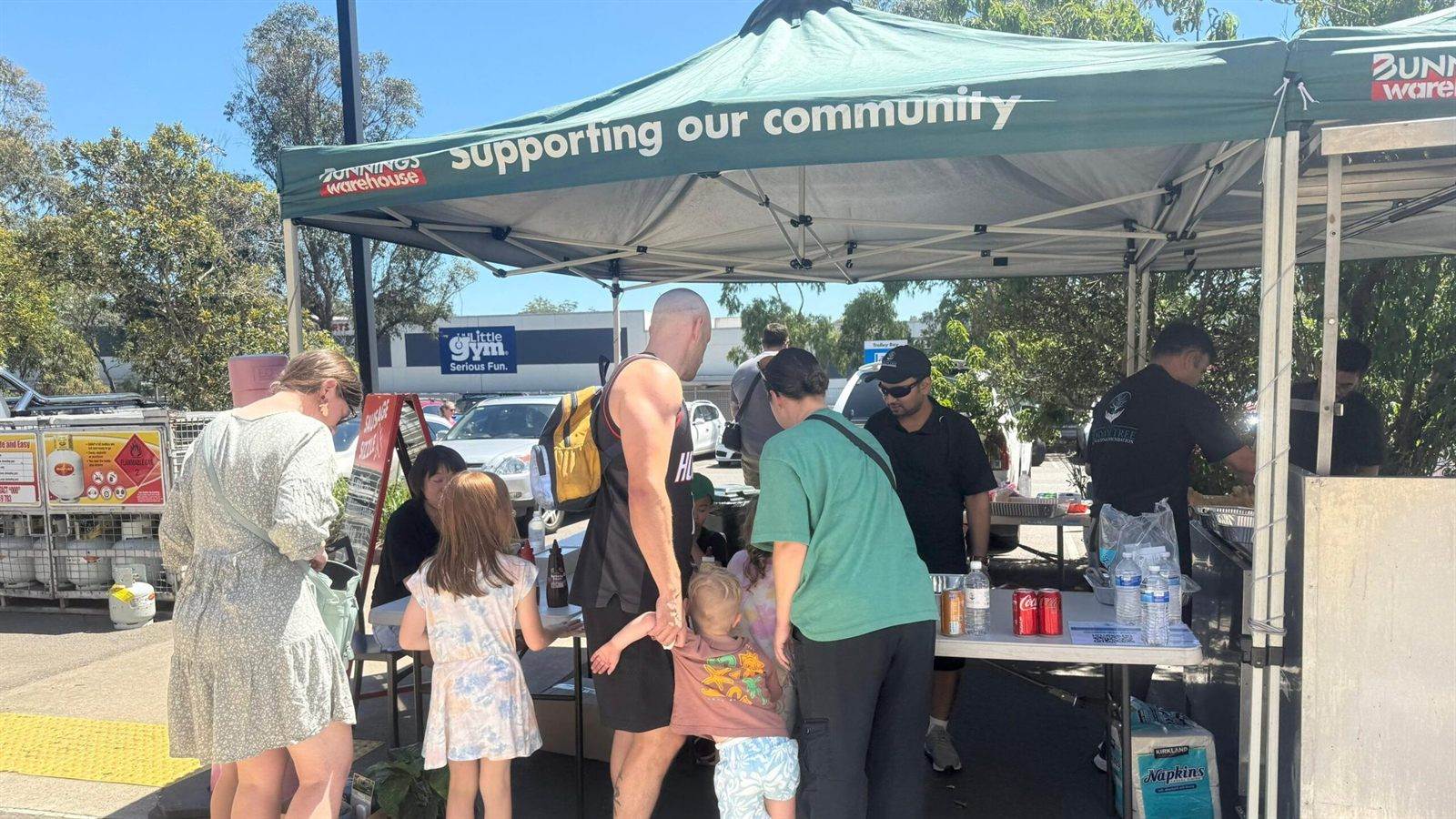 Parents and children waiting at a Bunnings barbecue while volunteers in black Wisdom Tree polos cook and serve