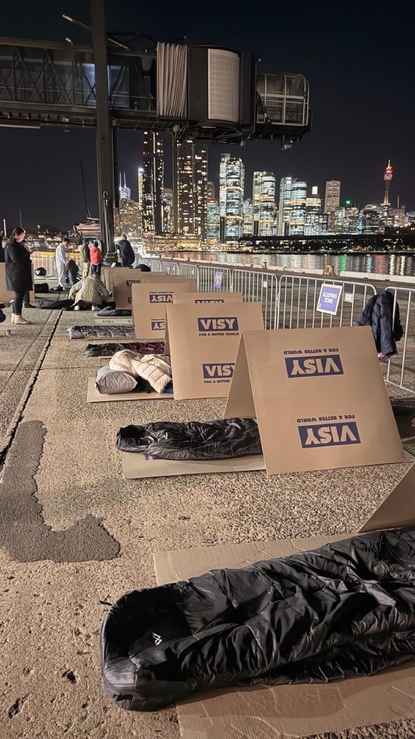 Cardboard shelters and sleeping bags laid out on concrete by the harbour, city towers lit behind