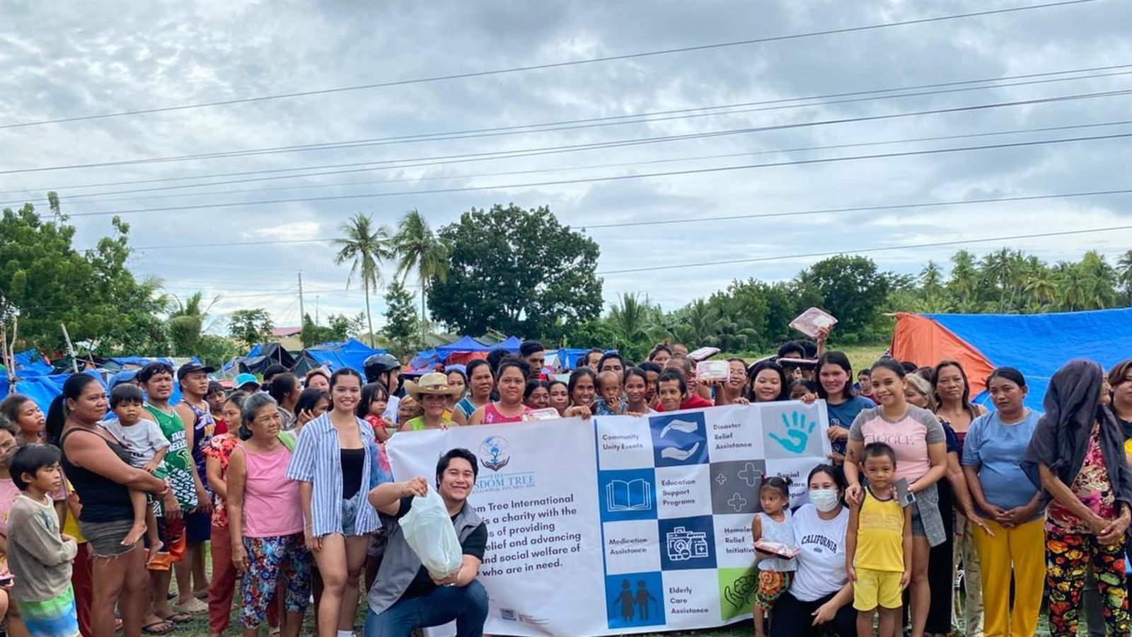 A large crowd of residents gathered in front of blue and orange tarpaulin shelters under a grey sky
