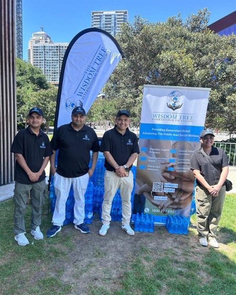 Four volunteers in Wisdom Tree caps and polos standing beside pallets of bottled water and a branded banner